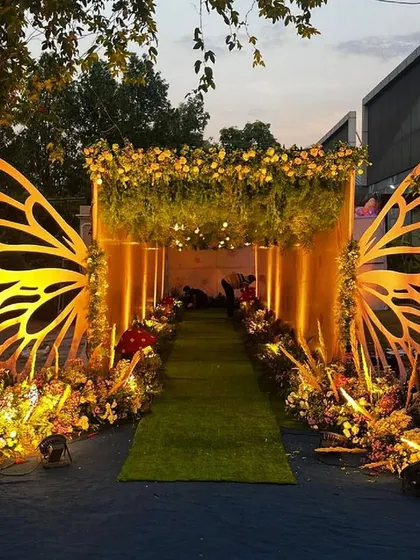 A whimsical entrance combining large butterfly wing props with a lush green walkway and warm uplighting. This setup is perfect for a fairytale or enchanted forest themed event.