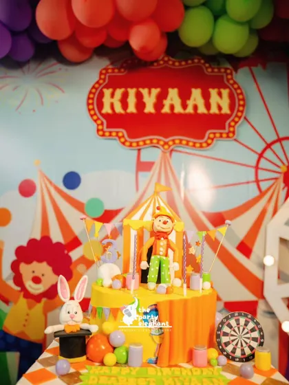The birthday cake is a centerpiece of the celebration. This custom carnival-themed cake features a clown, a magician's rabbit, and a dartboard, perfectly matching the party's decor and activities.