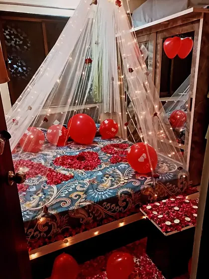 A cozy first-night room decoration with a canopy of fairy lights over the bed. The bed is covered in rose petals and red balloons to complete the romantic theme.
