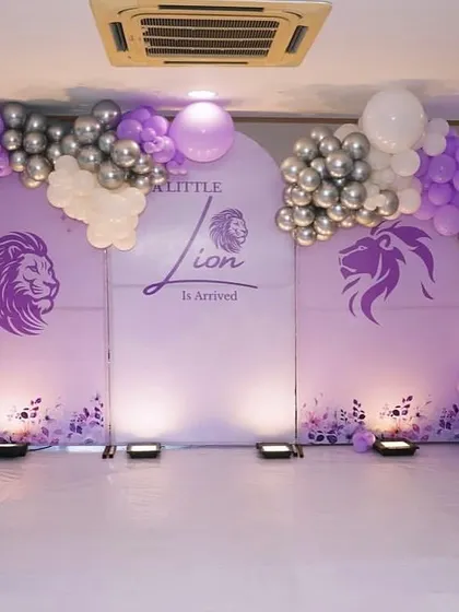 A "Little Lion Is Arrived" theme for a baby welcome or baby shower. The purple backdrop is decorated with a purple, white, and silver balloon arch and 'BABY' blocks.