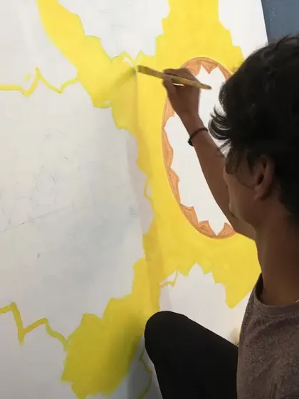 A close-up of an artist painting the base colors for a large mandala design on a canvas. This is the first step before the detailed clay and mirror work begins.