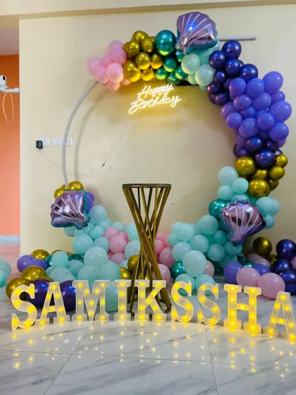 A mermaid-themed birthday decoration for a home party. The setup includes a circular balloon arch with metallic and pastel balloons, shell-shaped foil balloons, and marquee letters.