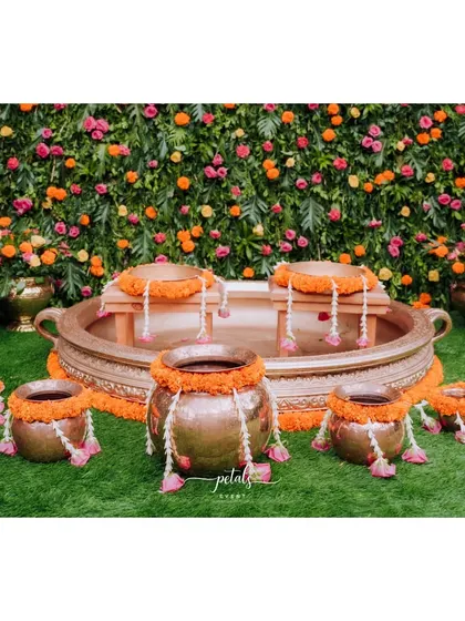 The Haldi setup features a large brass urli with traditional seating, set against a backdrop of a colorful flower wall. The combination of metallic elements and bright blooms is simply breathtaking.