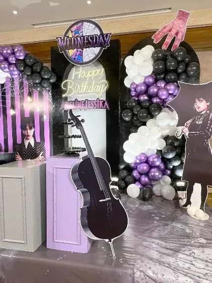 A side angle of the 'Wednesday' birthday party decor, highlighting the depth and various elements of the setup. This view shows the cello prop, character cutouts, and layered balloon arrangements that bring the theme to life.