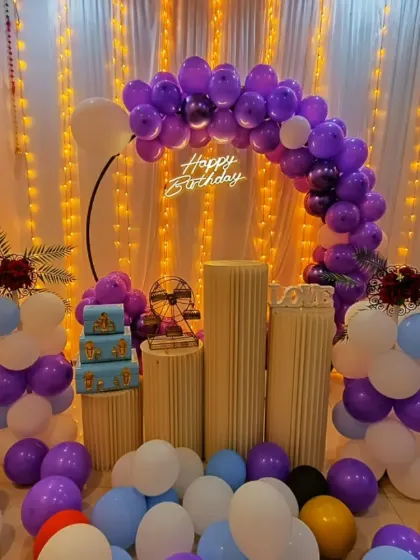 A birthday party setup with a purple and white balloon arch. The floor is filled with balloons, and the decor includes plinth stands and a mini Ferris wheel.