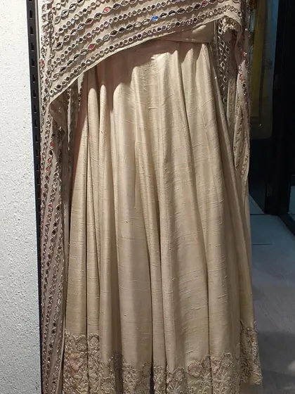 An elegant cream-colored Anarkali made from raw silk with a stunning cape-style dupatta featuring heavy mirror work. This is a luxurious and traditional piece for a major celebration.