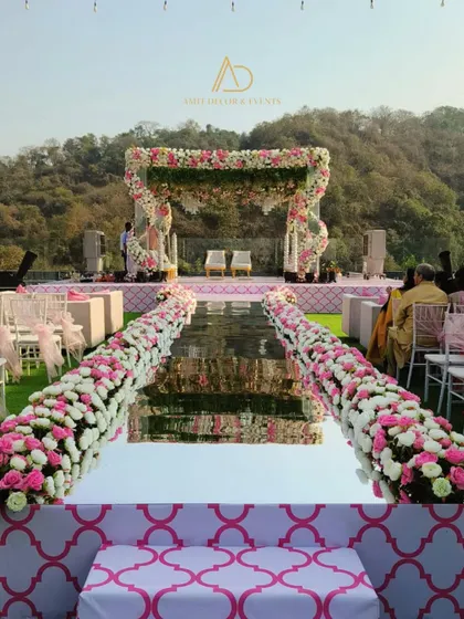 A mirrored aisle reflects the sky and the stunning floral mandap, creating a breathtaking path for the bride. The aisle is lined with dense arrangements of pink and white flowers.