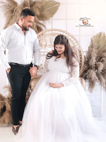 A beautiful couple's portrait from a bohemian-themed studio session. The partner holds his wife's hand as she sits gracefully in a white gown, surrounded by pampas grass.