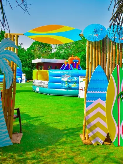 An entrance to the activity zone at the beach party, framed by bamboo poles and surfboard props. In the background, you can see a large inflatable water slide, perfect for outdoor fun.