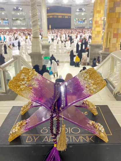 A beautiful photo of the purple and gold Quran holder in Makkah, with the Kaaba in the background, connecting the art to its spiritual source.