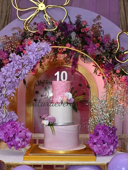 A grand cake display for an Aqeeqah and 10th birthday celebration. The beautiful tiered cake is framed by a golden arch and lush purple floral arrangements.