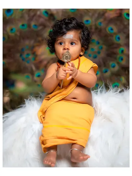 A full-body shot of the baby in the Lord Murugan costume, holding a miniature vel.