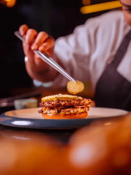 A chef carefully places a pickle on our Oklahoma-style smashed beef burger, the final touch on a culinary dream.