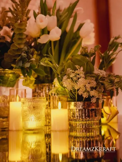 A close-up of a table centerpiece, featuring white tulips and other delicate blooms in gold vases, all reflected on a mirrored tabletop. The warm candlelight enhances the golden hues.