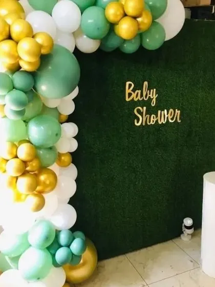 A beautiful baby shower backdrop using a faux grass wall. The wall is decorated with a 'Baby Shower' sign and a balloon garland in sage green, white, and gold.