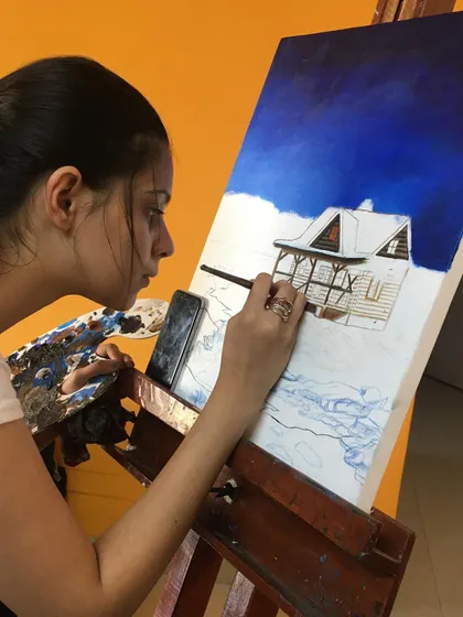 A student carefully works on her acrylic painting of a cozy, snow covered cabin. You can see her focus as she builds the scene, starting with the deep blue sky and working her way to the finer details of the house.