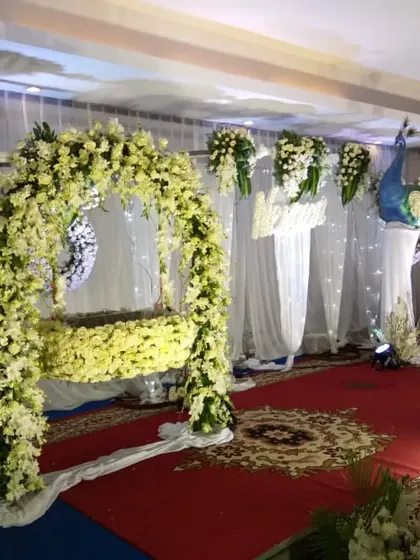 An elegant naming ceremony stage with a peacock theme. The decor features a fully decorated cradle, white floral arrangements, and a beautiful peacock statue with a tail made of flowers.