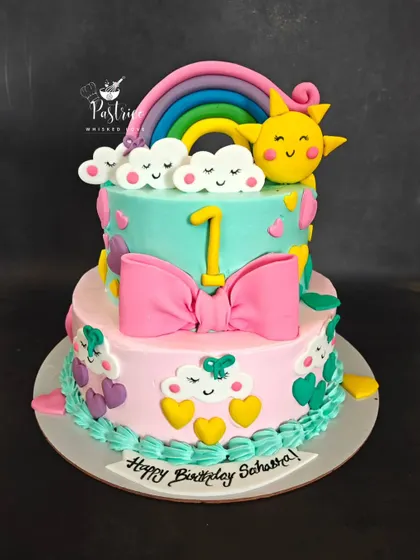 A cheerful two-tiered cake with a sunshine and rainbow theme, complete with cute clouds and a big pink bow.