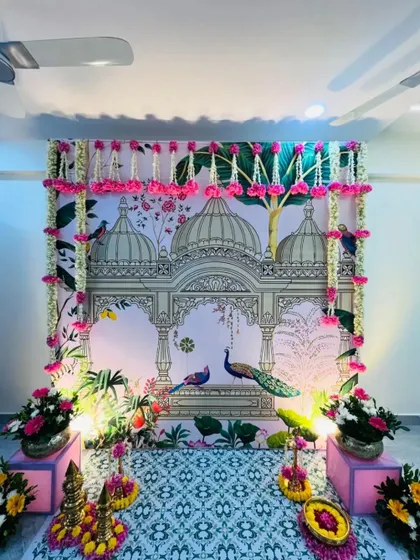 A stunning Pichwai backdrop used for a housewarming ceremony. The intricate architectural details and peacock motifs, framed by real flower garlands, create a grand and auspicious entrance.