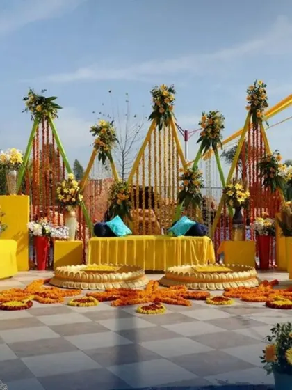 An expansive outdoor Haldi setup with modern triangular frames decorated with marigold strings. The open-air design includes multiple seating areas and traditional 'urli' bowls, perfect for a large gathering.