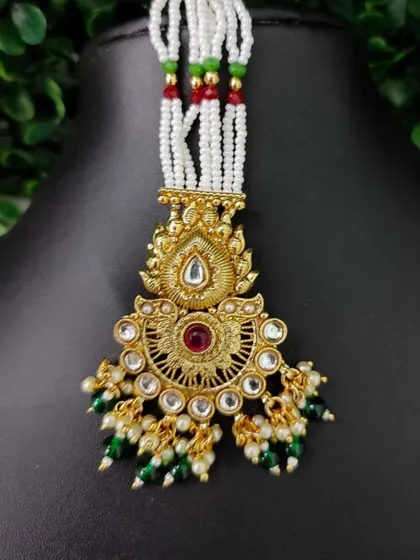 A temple-style maang tikka with a sunburst motif and a central red stone. The piece is attached to white pearl strands with red and green accents.