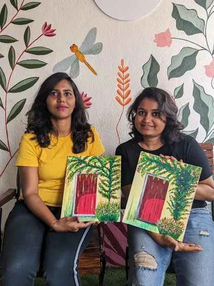 Two friends proudly holding up their finished canvas paintings of a red door surrounded by greenery. It's a joy to see people create together.