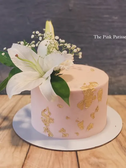 A simple yet stunning pink cake decorated with flecks of gold leaf. The design is topped with a single, elegant fresh white lily and a sprig of baby's breath.