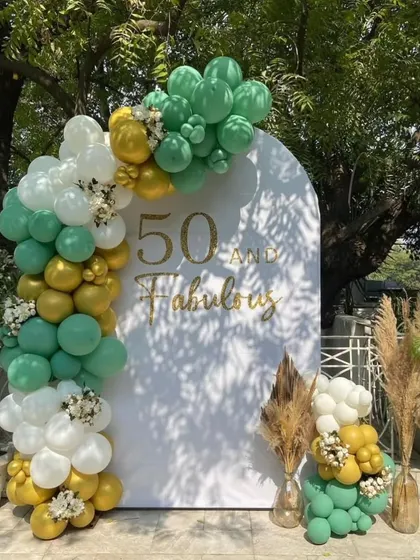 An elegant "50 and Fabulous" birthday celebration. This outdoor setup uses a custom white arched backdrop with a green, gold, and white organic balloon garland.