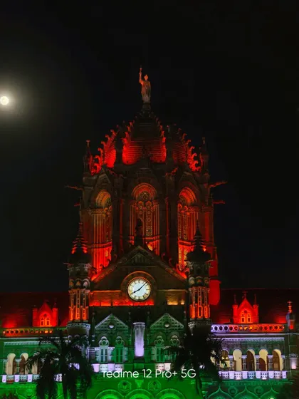 A crisp night shot of the Chhatrapati Shivaji Maharaj Terminus (CSMT) illuminated in the colors of the Indian flag, with the moon visible in the sky.