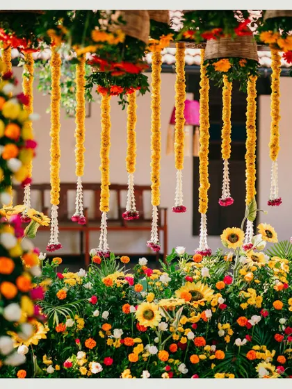 A close-up of the Haldi decor, showing the rich textures and colors of the floral arrangements. Hanging marigold strings and a backdrop of sunflowers and other vibrant blooms create a visually stunning setup.