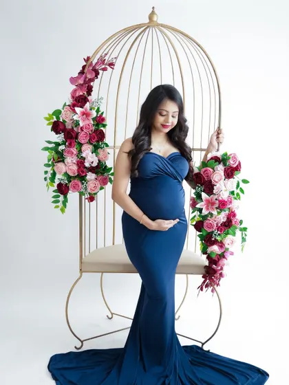 An elegant and classic portrait. The mom-to-be stands in a beautiful navy blue gown, holding her bump in a regal pose next to a floral birdcage prop.
