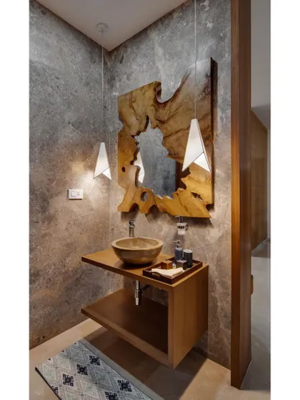 The powder room in the Nataraj House features a live-edge wood mirror and a stone-finish sink, set against a textured cement wall. This combination of raw, natural materials creates a grounded and sophisticated space.