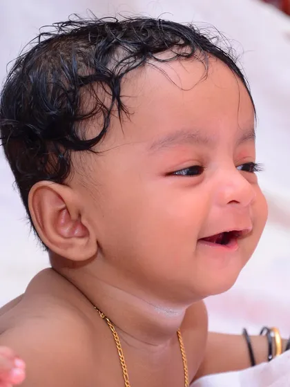 A side-profile candid shot of a smiling baby. This natural light portrait captures the simple joy and innocence of early babyhood, complete with traditional bangles.