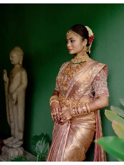 A bride, adorned in gold, stands next to a Buddha statue, a portrait that is both regal and serene.