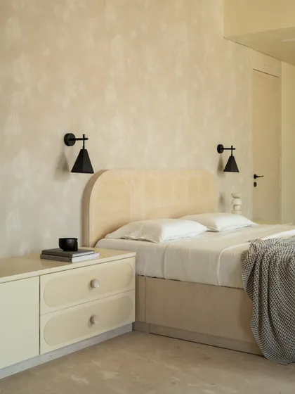 The bed in the Soft Serve master suite features a simple, curved headboard in a light veneer, set against a textured, lime-washed wall.