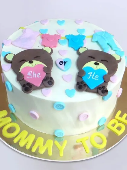 We create a sweet guessing game with this "Mommy to Be" gender reveal cake. Two teddy bears hold "She" and "He" hearts, surrounded by a clothesline of baby outfits, making for an adorable and interactive baby shower centerpiece.