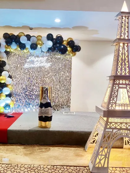 A Paris-themed birthday party featuring a large light-up Eiffel Tower prop. The backdrop includes a silver shimmer wall and a black and gold balloon arch.
