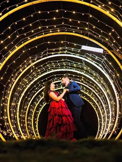 A romantic moment in a beautifully lit tunnel of lights. She is wearing a tiered red ruffle gown, and he is in a blue suit, creating a magical and memorable photo.