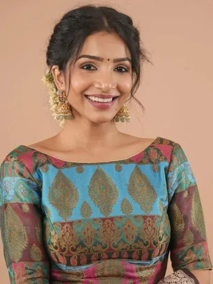 A smiling model wears a colorful brocade blouse with a boat-neck design, paired with traditional gold jhumkas.