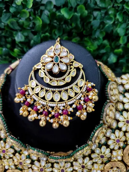 A close-up of a maang tikka from a rental set, featuring a crescent design with kundan work and ruby-colored bead drops.
