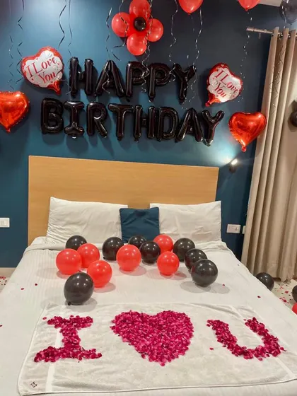 An intimate room decoration for a birthday. The setup includes "Happy Birthday" foil balloons, heart-shaped balloons, and an "I Love You" message written with rose petals on the bed.