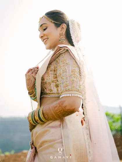 A beautiful outdoor shot of our bride Poojita, looking ethereal in her pastel pink silk saree. The sunlight catches the intricate gold and pearl embroidery on her custom blouse, highlighting the temple-inspired motifs on the sleeves.