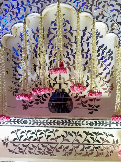 Intricate details on a stage backdrop. A mirrored mosaic in a floral pattern is adorned with hanging strings of jasmine and pink flowers, creating a delicate and beautiful effect.