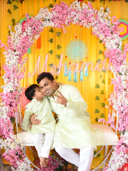A full-length shot of the father and son in matching outfits, posing against the festive Mehendi backdrop.