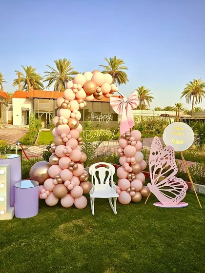 A beautiful butterfly-themed first birthday in a garden. This setup features a pink and gold balloon arch, a large butterfly cutout, and a neon 'Happy Birthday' sign.