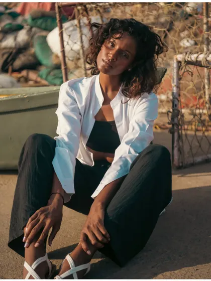 A relaxed, seated portrait in a rustic, industrial beach setting. The warm sunlight gives the shot a natural and approachable feel.