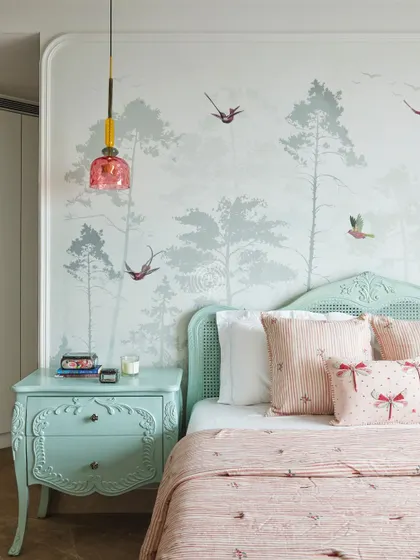 This girl's bedroom is soft and dreamy, with pastel green furniture and a delicate wallpaper featuring birds and trees. The pink glass pendant light adds a touch of color and whimsy.