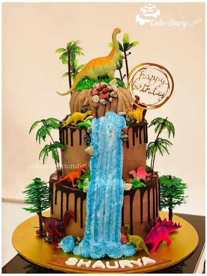 A magnificent dinosaur-themed cake with a waterfall. This multi-tier chocolate drip cake is decorated with various dinosaurs, palm trees, and a flowing blue waterfall made of buttercream.