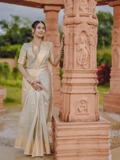 A full-length portrait of a bride in a traditional white and gold saree, set against ancient carved pillars.