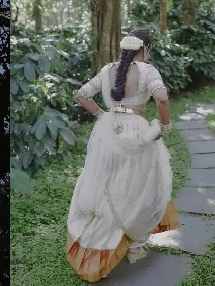 A candid shot of a bride in a white and gold half-saree, walking through a garden. This photo captures a moment of grace and movement, telling a story of new beginnings.